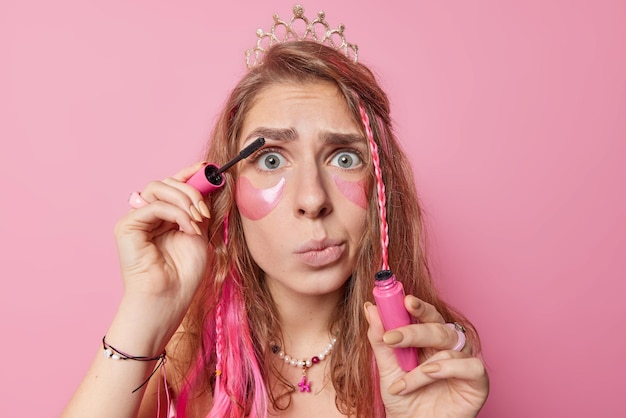 Portrait of surprised European woman with long hair applies mascara and beauty pads wants to look beautiful during birthday celebration wears crown on head purses lips poses against pink background