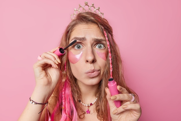 Free photo portrait of surprised european woman with long hair applies mascara and beauty pads wants to look beautiful during birthday celebration wears crown on head purses lips poses against pink background