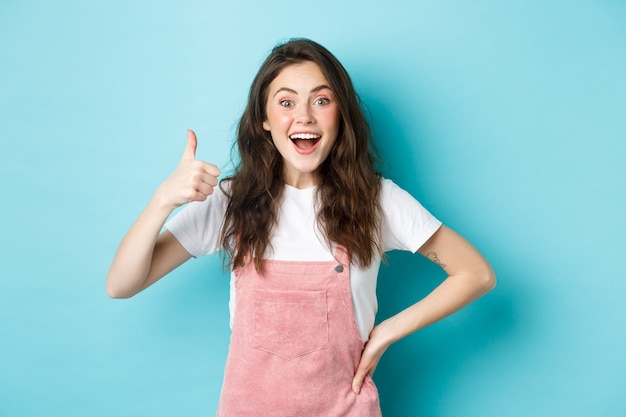 Portrait of surprised and excited brunette girl showing thumb up and say yes, gasping fascinated, praising great job, standing against blue background.