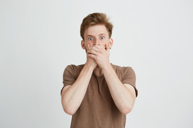 Portrait of surprised frightened scared young man closing mouth with hands.