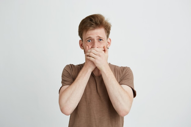 Portrait of surprised frightened scared young man closing mouth with hands.