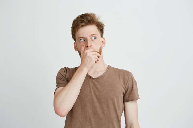 Portrait of surprised frightened scared young man looking in side closing mouth with hands.