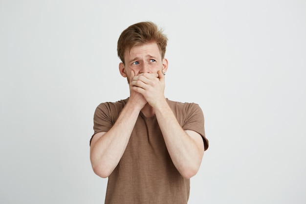 Portrait of surprised frightened scared young man looking in side closing mouth with hands.