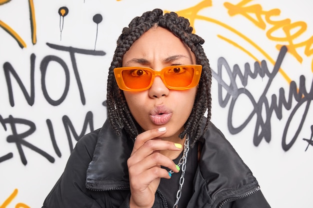 Portrait of surprised funky female teenager keeps hand on chin looks with wondered expression at camera wears orange sunglasses black jacket poses against painted graffiti wall