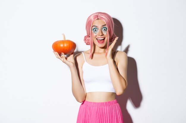 Portrait of surprised girl in pink wig, holding pumpkin and looking excited, celebrating halloween, standing.