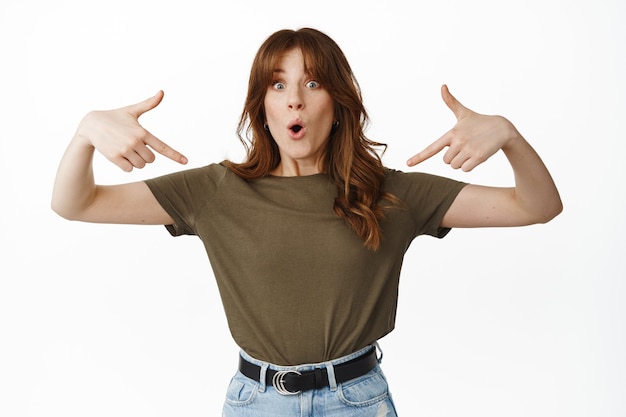 Free photo portrait of surprised girl student pointing fingers down at center and amazed standing on white