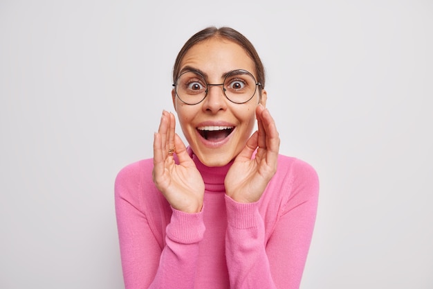 Portrait of surprised good looking young woan stares with great wonder at camera gasps from shock wears round spectacles pink turtleneck on white