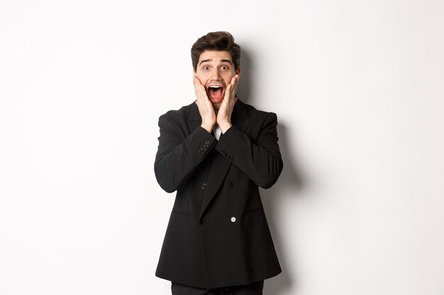 Portrait of surprised handsome businessman in suit, reacting to holidays promo, looking amazed at camera, standing over white background