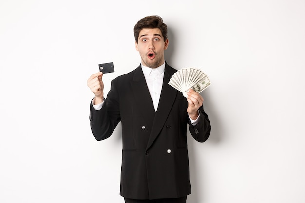 Portrait of surprised handsome man i suit, showing credit card with money, standing against white background