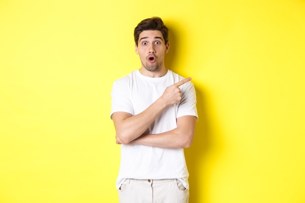 Portrait of surprised handsome man pointing finger right, gasping amazed, standing over yellow background.