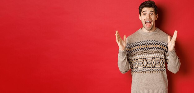 Portrait of surprised and happy handsome man wearing christmas sweater looking amazed celebrating new year standing over red background