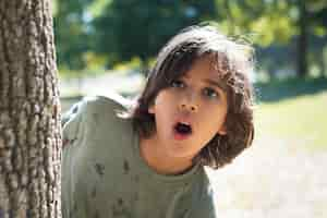 Foto gratuita ritratto del ragazzino sorpreso nel parco. ragazzo dai capelli scuri che dà una occhiata fuori dall'albero, guardando la fotocamera. famiglia, amore, concetto di infanzia