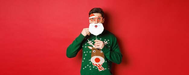 Portrait of surprised man in green christmas sweater, holding funny santa claus mask, celebrating new year, standing over red background.