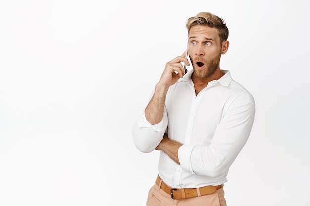 驚いた男の肖像画は、白い背景の上に立っているスマートフォンで電話をかけている携帯電話で面白いsmthを聞きます