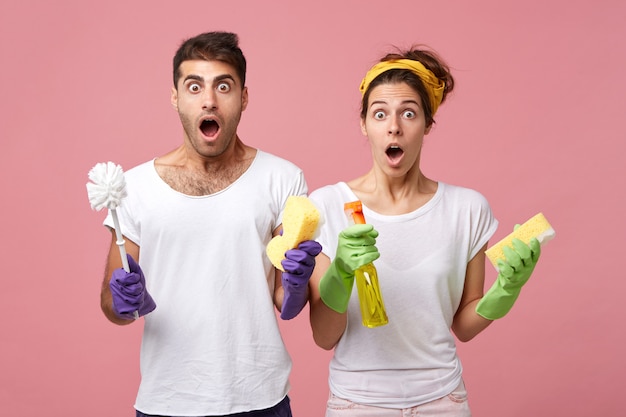 Portrait of surprised man wearing protective gloves holding brush and woman with sponge and detergent looking with widely opened eyes and mouths