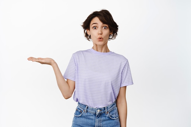 Portrait of surprised modern girl brunette woman holding smth empty open hand showing product on palm wow face receive something interesting white background