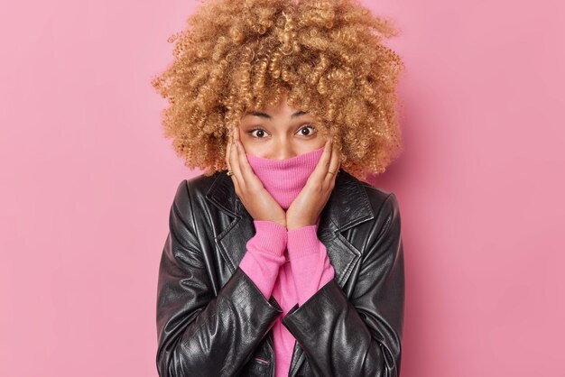 Portrait of surprised positive young woman with curly hair covers mouth with collar dressed in leather jacket looks directly at camera poses against pink background. People and emotions concept