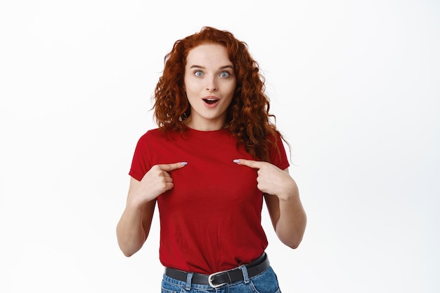 Portrait of surprised redhead female model pointing at herself and gasping from disbelief being chosen or winner smiling happy white background