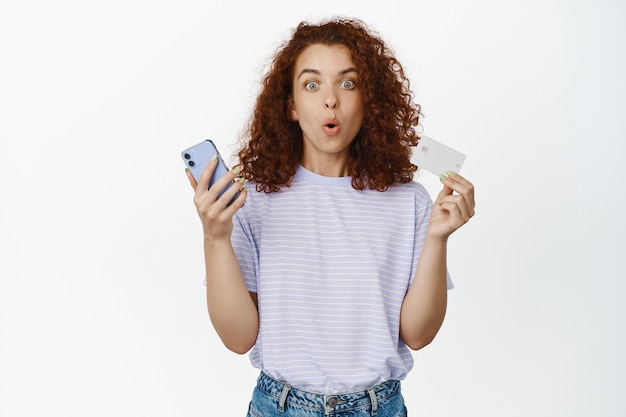 Portrait of surprised redhead girl shows her smartphone and credit card, amazed with online sale, discounts on application, standing on white