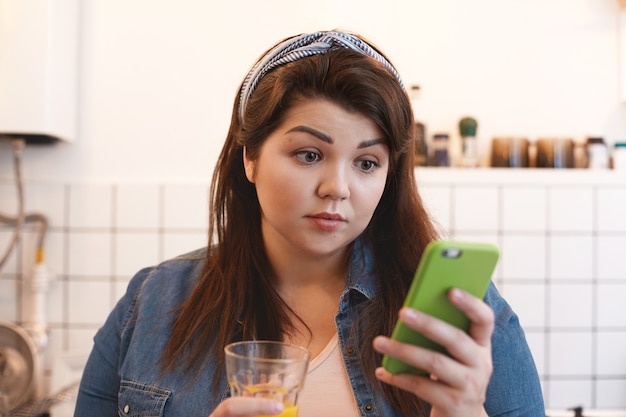 スマートフォンを使用して自家製の柑橘類のデトックスジュースを飲んで、予期しないニュースに驚いて驚いたショックを受けた若いぽっちゃり女性の肖像画。人、健康的なライフスタイルと技術の概念