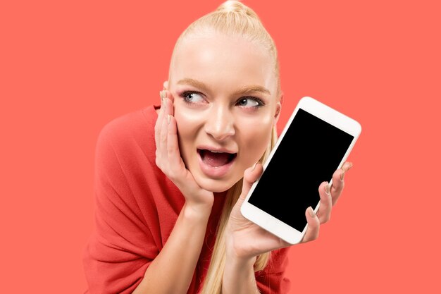 Portrait of a surprised, smiling, happy, astonished girl showing blank screen mobile phone isolated over coral background.