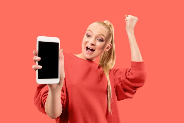 Portrait of a surprised, smiling, happy, astonished girl showing blank screen mobile phone isolated over coral background.