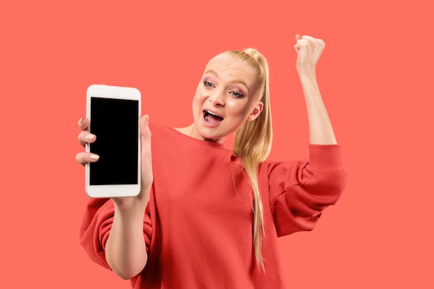 Портрет удивленной, улыбающейся, счастливой, удивленной девушки, показывающей пустой экран мобильного телефона, изолированного на коралловом фоне.