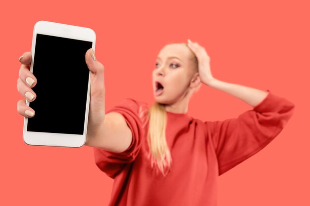 Portrait of a surprised, smiling, happy, astonished girl showing blank screen mobile phone isolated over coral wall