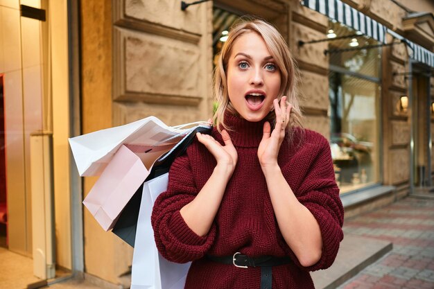 街の通りのカメラで驚いて見ている買い物袋を持つ驚いたスタイリッシュなブロンドの女の子の肖像画