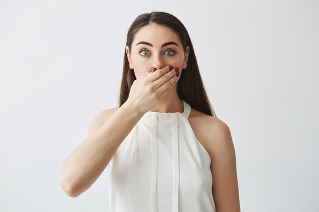 Portrait of surprised young brunette girl covering mouth with hand .