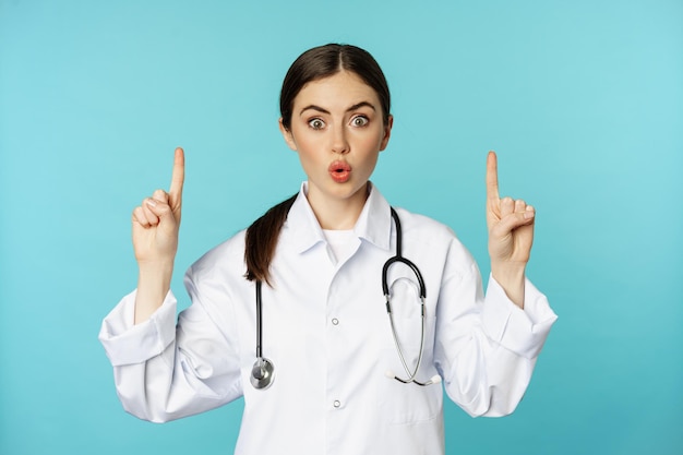 Portrait of surprised young doctor female medical worker pointing fingers up gasping amazed showing ...