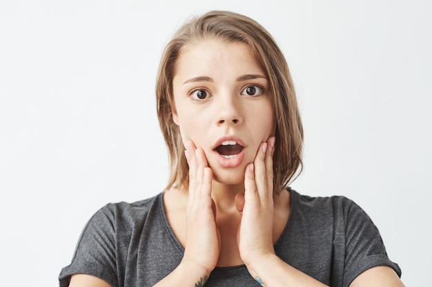 Portrait of surprised young pretty girl with opened mouth holding cheeks .