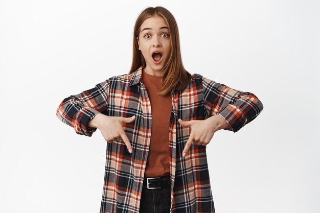 Portrait of surprised young woman gasp in awe, checking out big news, discounts and prices below, pointing down amazed, standing against white background