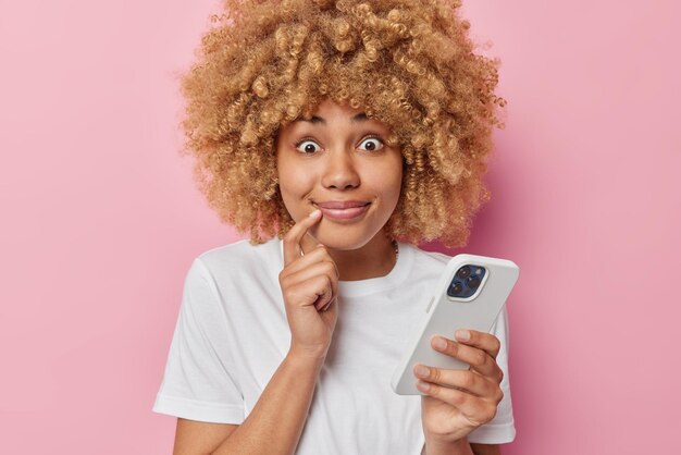 Portrait of surprised young woman keeps finger near lips uses mobile phone gets shocking news wears casual white t shirt isolated over pink background People reactions and technology concept