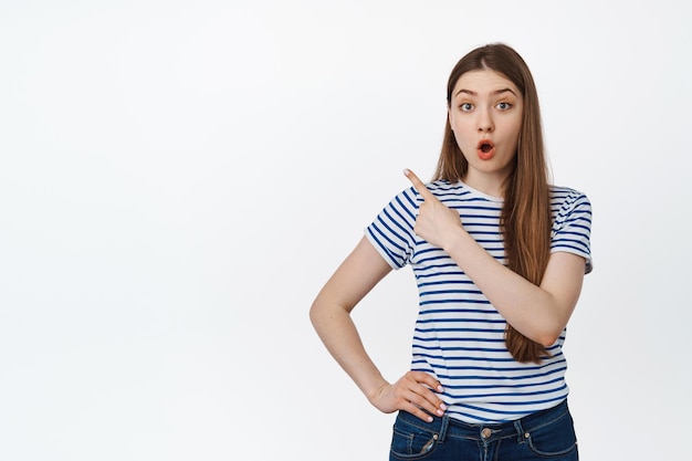 Free photo portrait of surprised young woman pointing at upper left corner, showing exciting announcement, standing over white background
