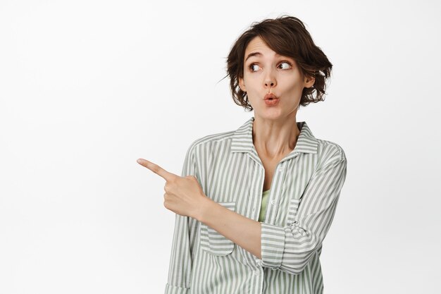 Portrait of surprised young woman pucker lips curious, pointing and looking left standing on white