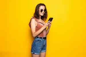 Free photo portrait of a surprised young woman in summer hat and sunglasses using mobile phone isolated over yellow wall