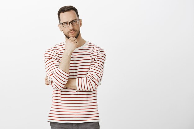 Portrait of suspicious thinking attractive man in striped clothes and glasses, looking aside and frowning, touching bristle, being concerned with problematic decision