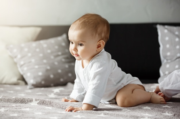 Free photo portrait of sweet  baby sitting on cozy bed. child looking aside and happily crawls to mother. family, motherhood concept.