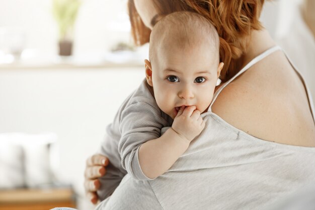 Portrait of sweet little child with big grey eyes and putting hand in mouth on mother shoulder. Mom hug her child with love. Family concept.