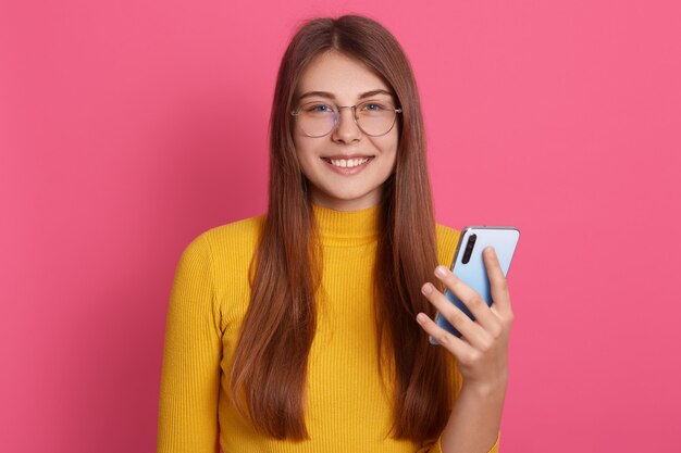 Portrait of sweet sincere adorable attractive young female having pleasant smile, holding smartphone, being in good mood, standing isolated over pink wall. People and technology concept.