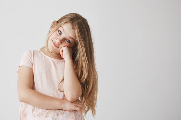 Portrait of sweet small girl with light long hair dressed in pink t-shirt  with joyful look, holding head with hand.