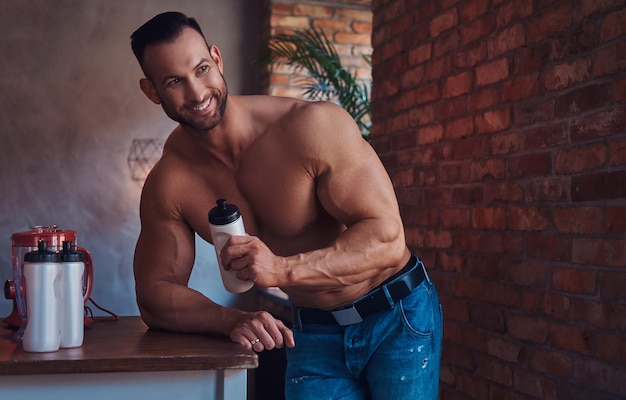 Portrait of a Tall stylish shirtless bodybuilder dressed in jeans, prepares protein mixture before morning exercise, standing in a kitchen with a loft interior, looking out the window.