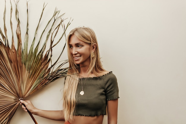 Portrait of tanned blonde long-haired woman in dark green t-shirt smiles sincerely and holds dry palm leaf on white background