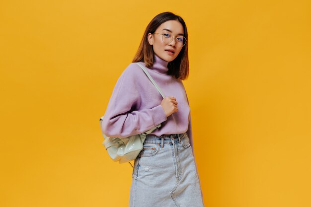 Portrait of tanned short-haired young woman in eyeglasses, denim skirt and purple sweater looks at front and holds mint backpack on orange wall