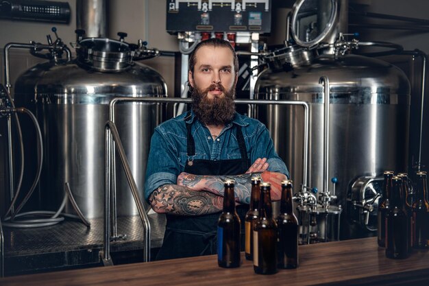 Portrait of tattooed, bearded hipster male manufacturer presenting beer in the microbrewery.