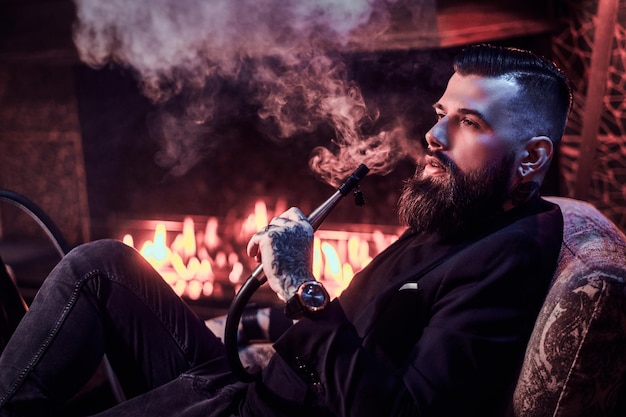 Portrait of tattooed bearded man which is smoking hookah, making nice vapour while chilling on the armchair near fire place.