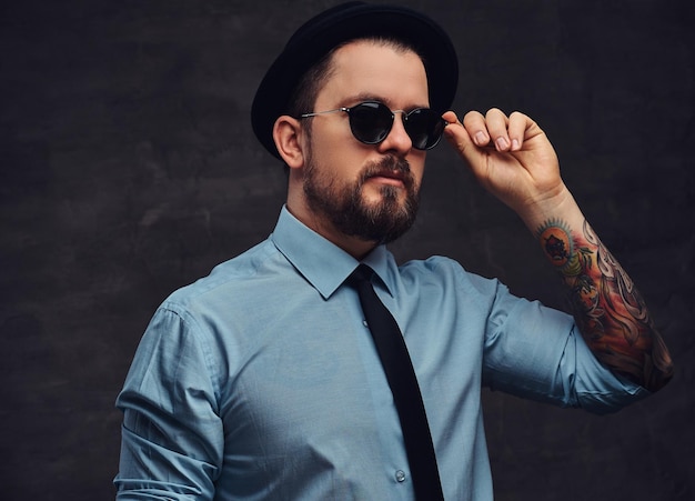 Portrait of a tattooed handsome middle-aged man with beard and hairstyle dressed in formal clothes and hat, pose with hand on sunglasses. Isolated on a textured dark background in a studio.