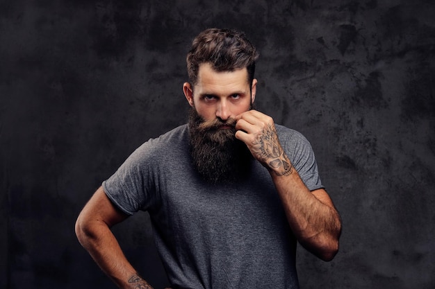 Portrait of a tattooed hipster with full beard and stylish haircut, dressed in a gray t-shirt, looking at the camera, stands in a studio on a dark background.