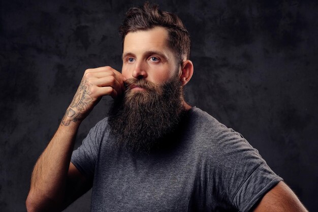 Portrait of a tattooed hipster with full beard and stylish haircut, dressed in a gray t-shirt, stands with a thinking look in a studio on a dark background.